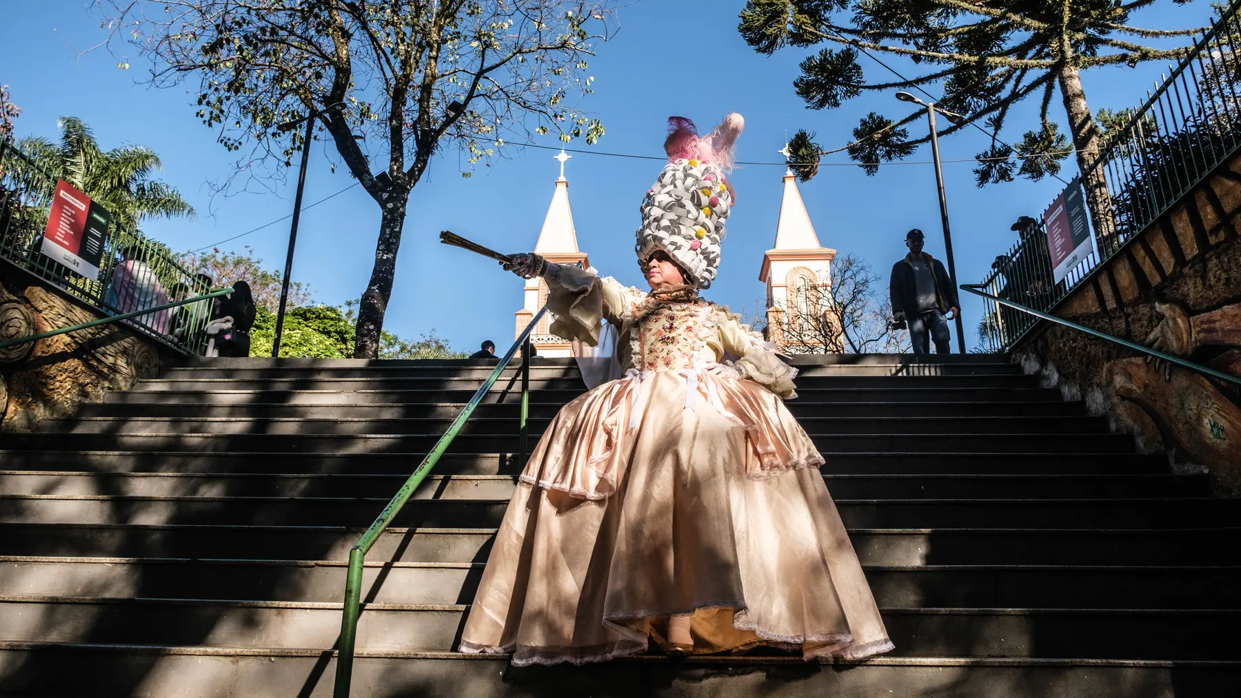 Drag em vestido de época e peruca monumental desce as escadarias da praça, com as torres da catedral ao fundo.