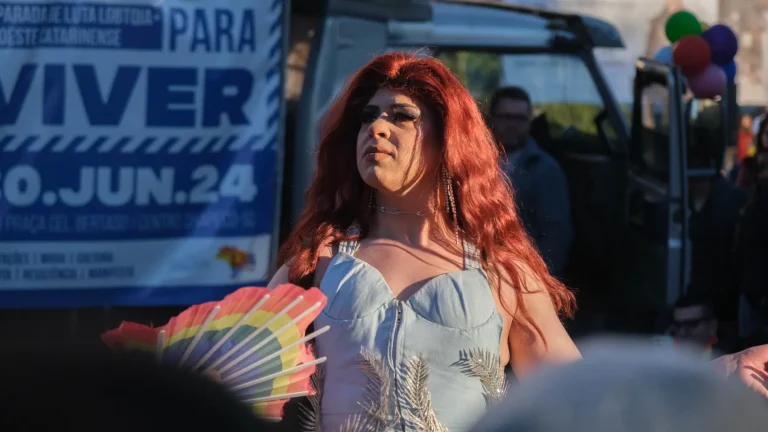 Red-wigged drag queen with a rainbow fan in front of the “Para viver — 30.jun.24” banner.