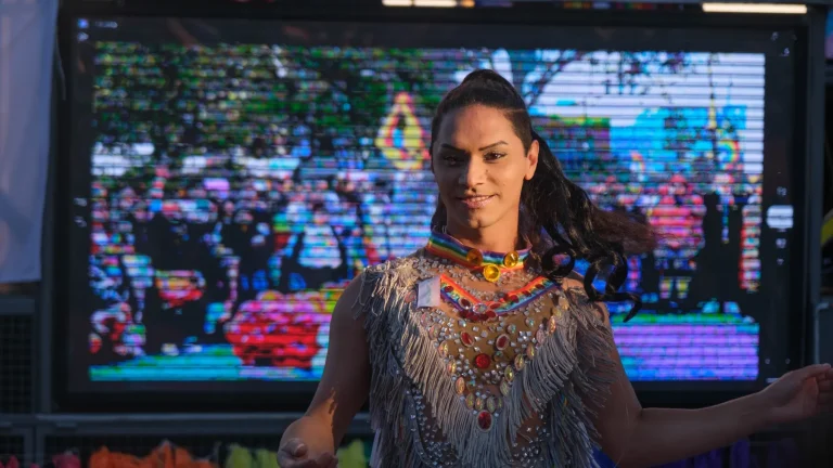 A performer in fringes and colorful necklaces dances before the float’s screen.