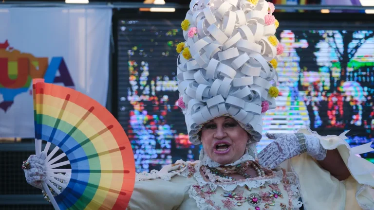 The sculpted-wig drag queen opens her rainbow fan atop the float.