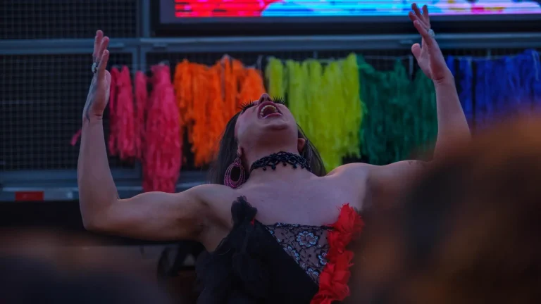 Arms up: a drag queen in ecstasy against rainbow fringes.