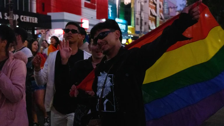 A pride flag spread across the avenue as shop lights come on.
