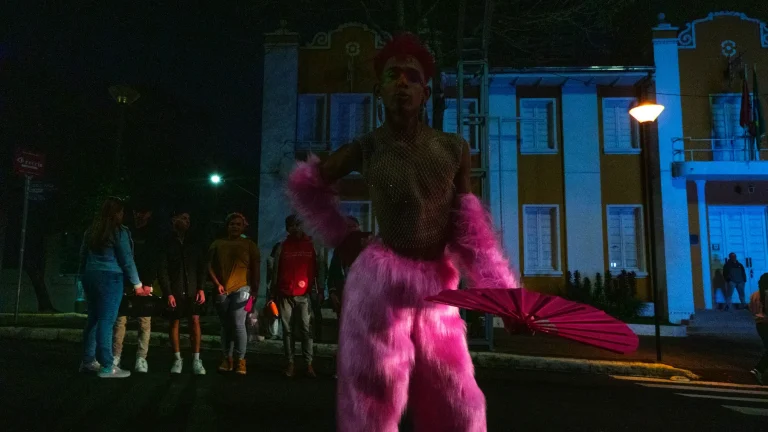 A drag queen in pink feathers dances in the street at night.
