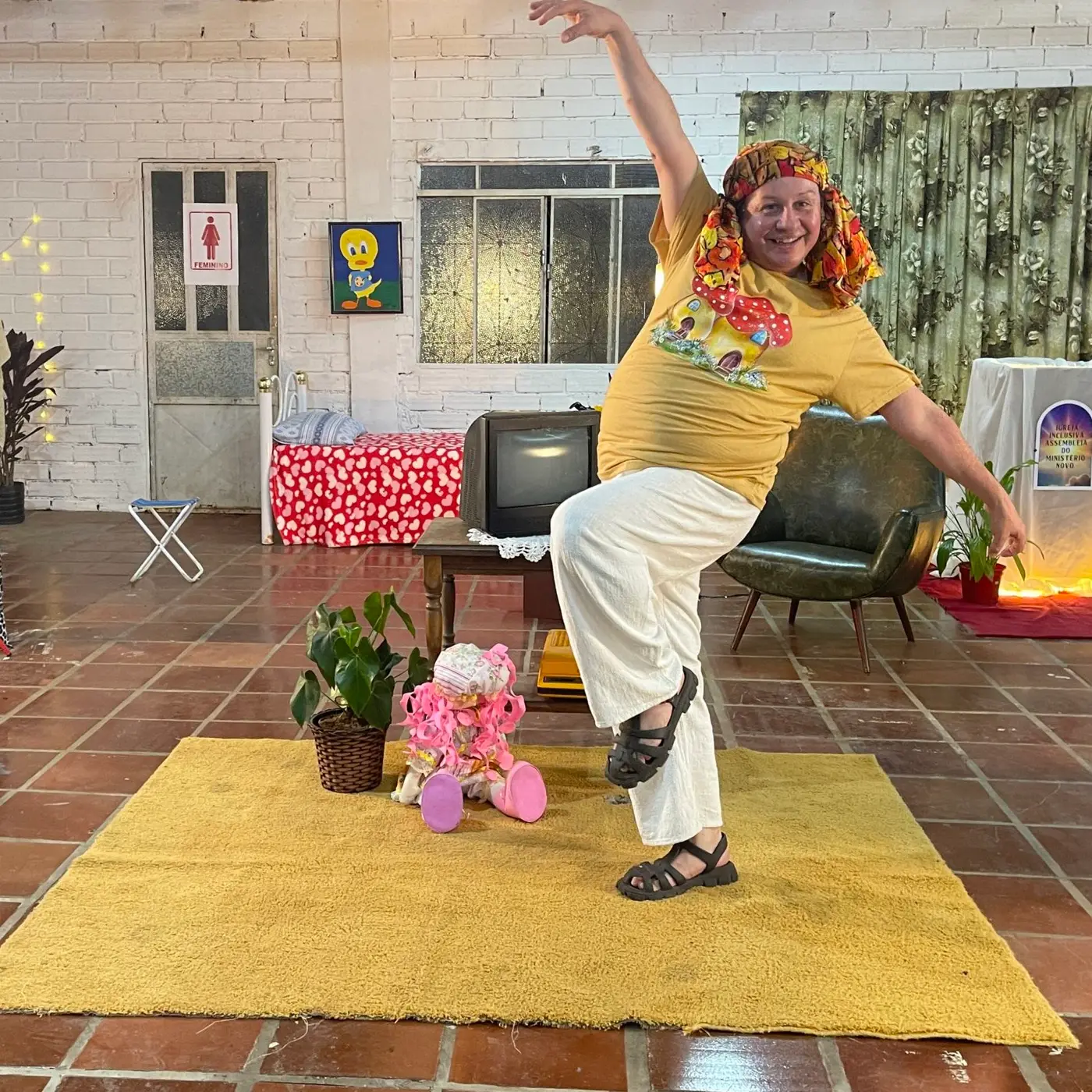 Performer dança de braço erguido no cenário da videoinstalação Vem de Berço, com tapete amarelo, TV antiga e poltrona