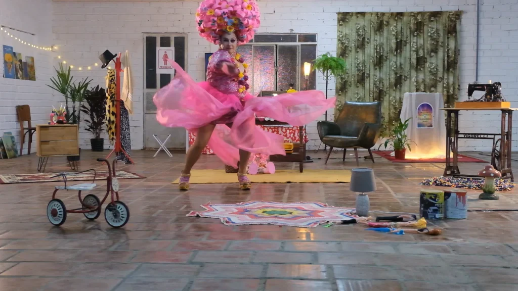 Vem de Berço: uma drag queen de vestido rosa e cabelo de flores rodopia no cenário de memórias de infância